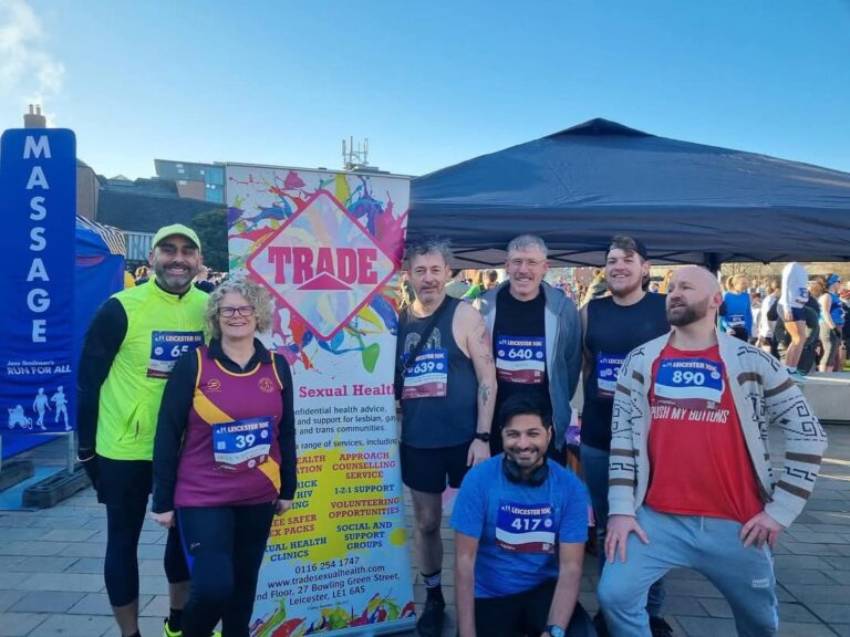 A group of happy and excited people, running the Leicester 10k Run for All in support of Trade, stand in front of a promotional banner.
