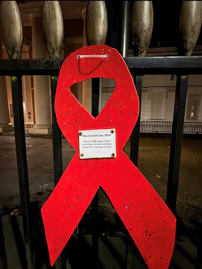 A large wooden red AIDS ribbon attached to fencing. Writing in the centre says 'World AIDS Day 2024. Nearly 10,000 people visited the Positive Leicester exhibition about HIV in Leicester in 2022'.