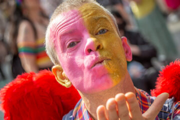 Person with face painted half pink and half gold glitter blows a kiss to the camera.