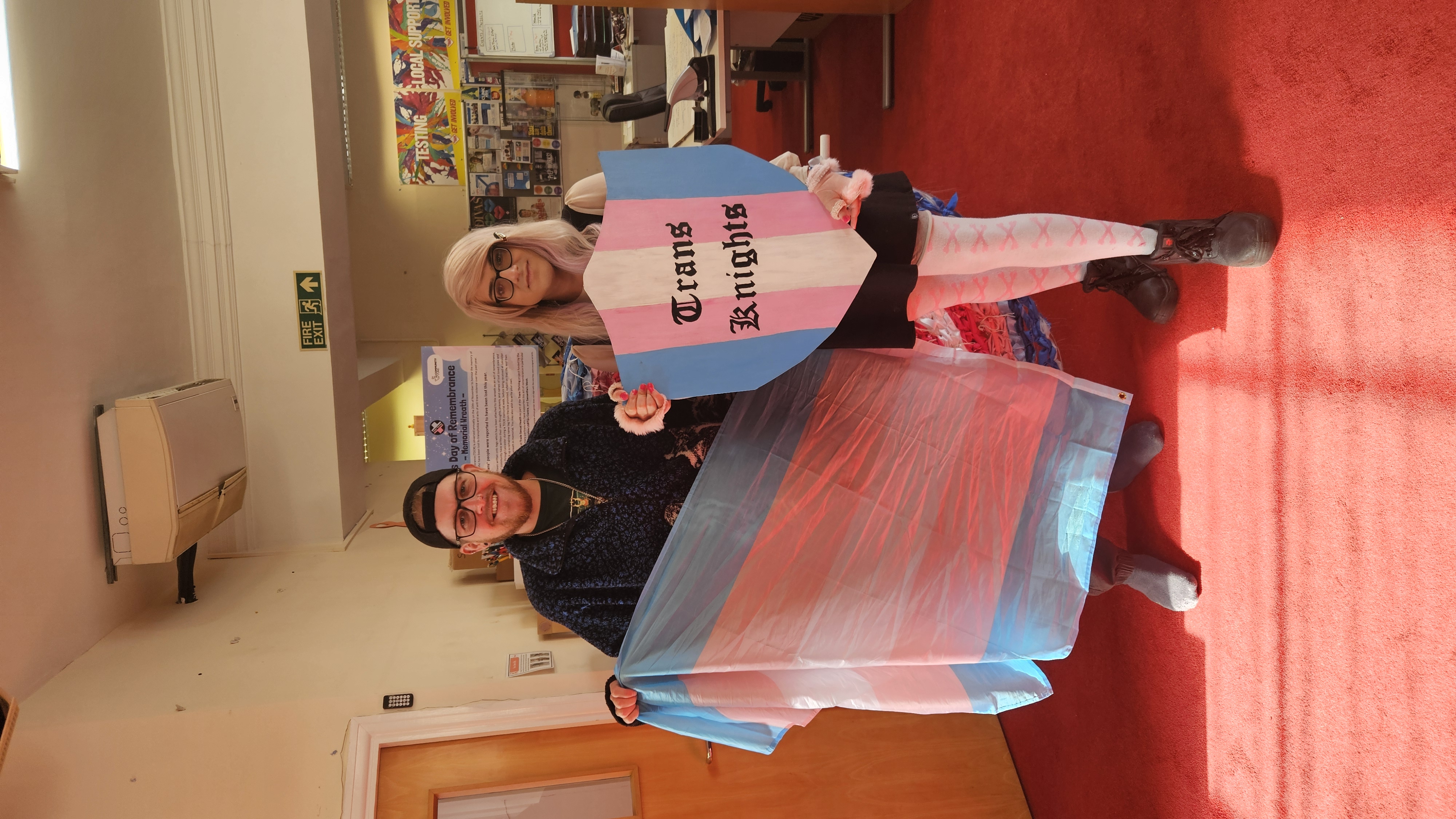 Two people pose with a Trans flag and a shield that says 'Trans Knights' in the Trans flag colours.